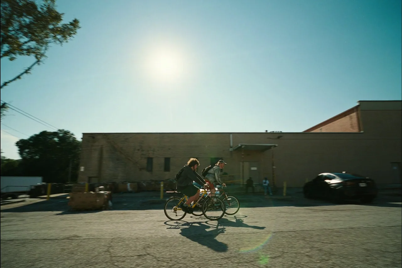 Bugonia (2025) movie still: wide — An asphalt parking lot sits before a low beige brick industrial building. Two young men ride bicycles across the…