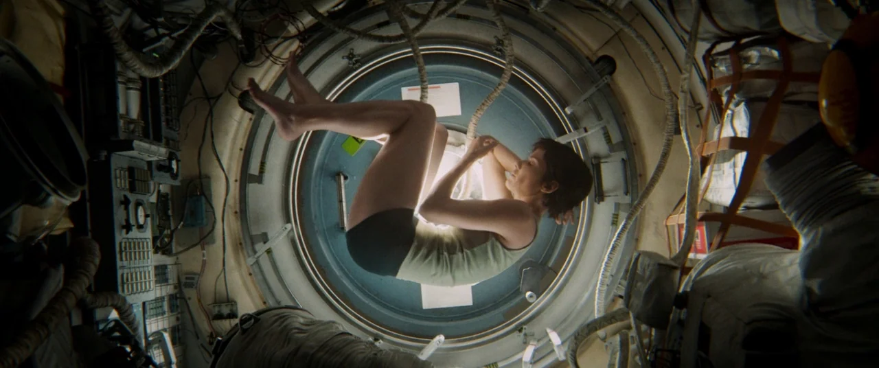 Gravity (2013) movie still: fullbody — The interior of a cramped spacecraft module features metallic walls covered in exposed cables. A woman floats…