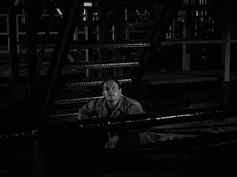 White Heat (1949) movie still: medium — A dark industrial space containing metal stairs and structural beams. The man crouches on the floor beneath…