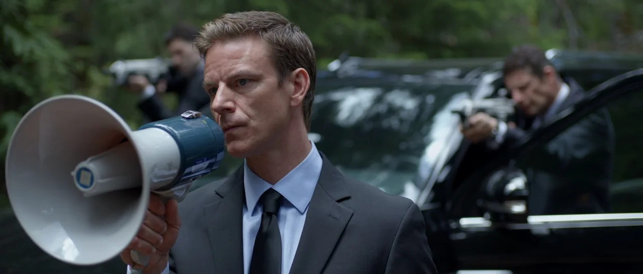 La presencia (2012) movie still: medium — A man in a suit stands in a forest holding a megaphone near a vehicle. Two men holding firearms exit a…