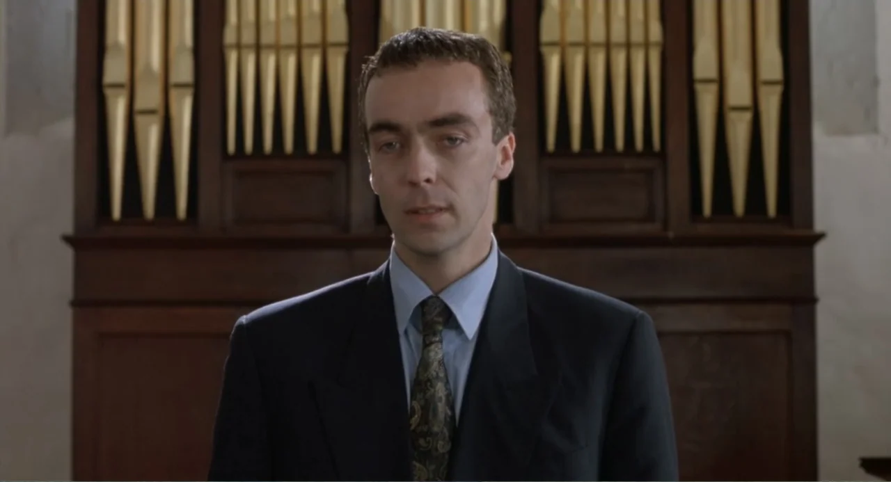 Four Weddings and a Funeral (1994) movie still: medium — A man stands in front of large wooden organ pipes in a church. He is wearing a dark suit…