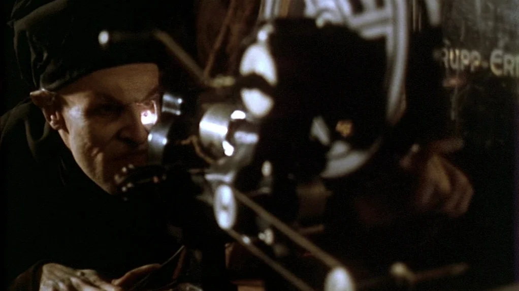 Shadow of the Vampire (2000) movie still: medium — A dark, cramped space contains a vintage metal film projector with large mechanical reels. A bald…