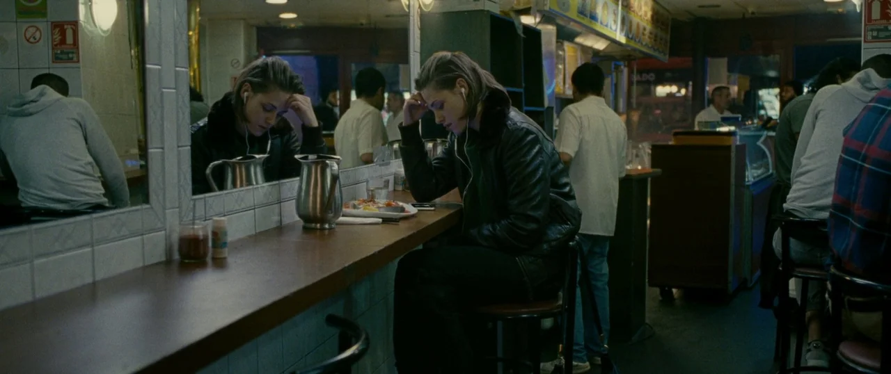 Personal Shopper (2016) movie still: medium — A dimly lit diner with tiled walls and a mirror behind the counter. A young woman sits at the counter…
