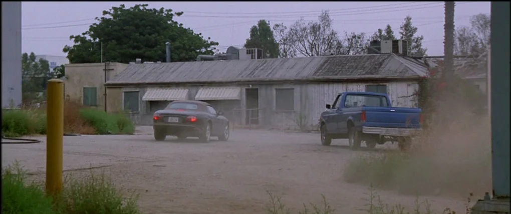 Memento (2000) movie still: wide — A dusty lot with a single-story white building stands in a rural area. A blue truck and a dark car are positioned…