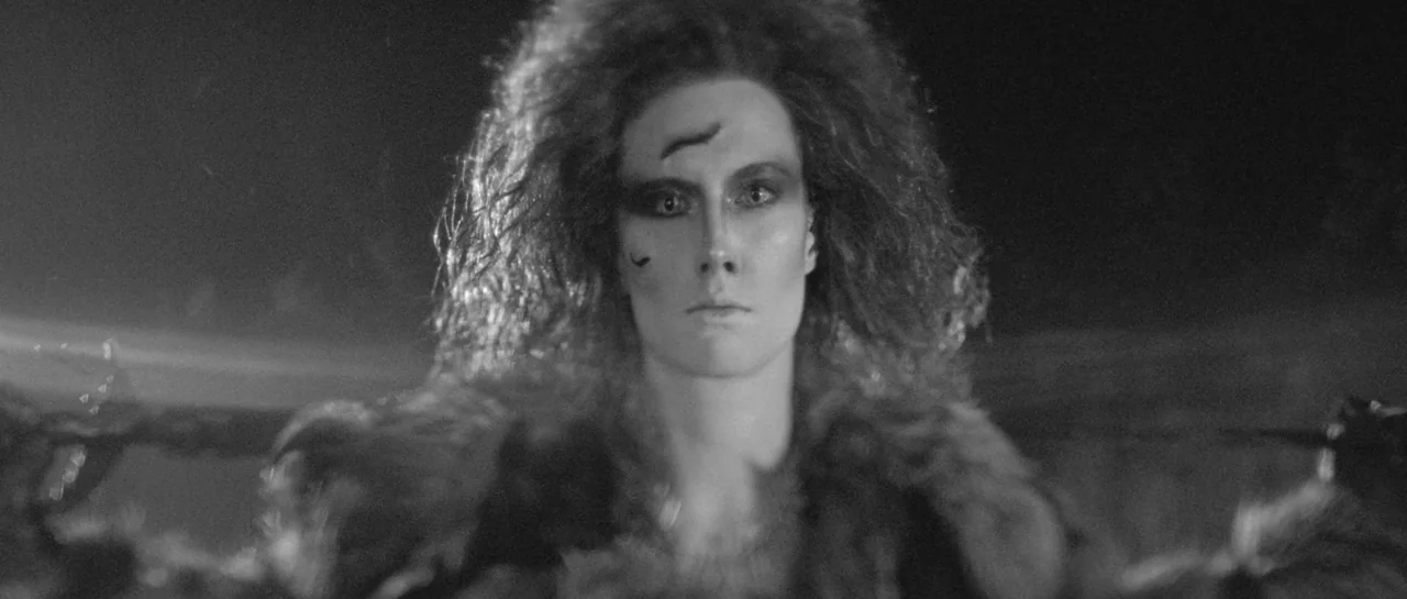 She Is Conann (2023) movie still: closeup — A person with wild hair and dark markings stands in a dark outdoor environment. They face forward toward…