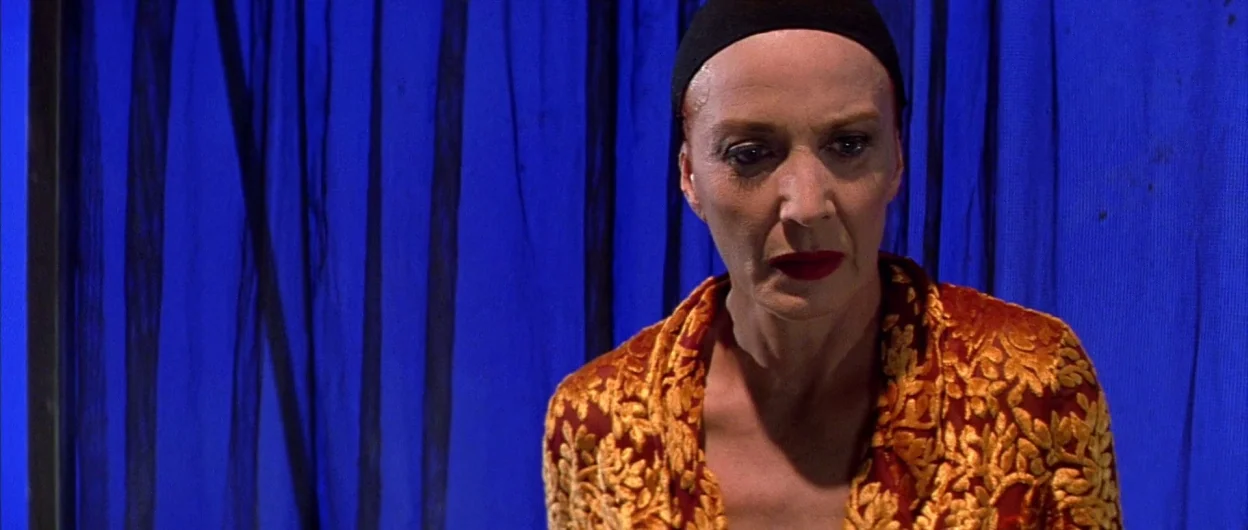 All About My Mother (1999) movie still: medium — A woman stands before a vibrant blue curtain stretched tightly against a frame. The subject wears a…