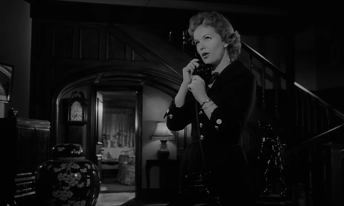 Night Of The Demon (1957) movie still: medium — A dimly lit 1950s home foyer features dark wood paneling and a grand staircase. A woman stands in…