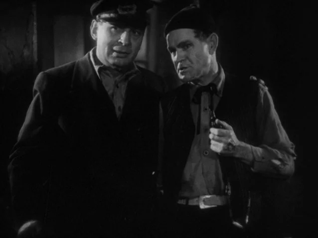 The Docks of New York (1928) movie still: medium — A dark interior space with minimal architectural detail visible in the low light. Two men stand…