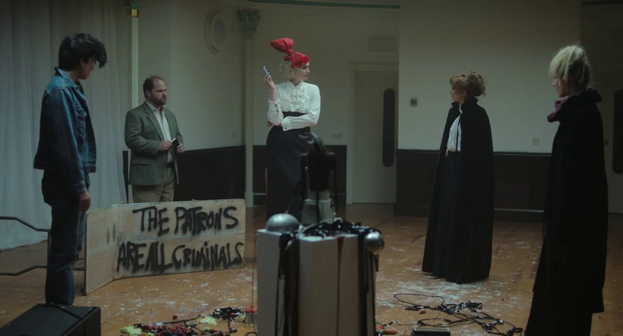 Flux Gourmet (2022) movie still: wide — A spacious room features hardwood floors littered with debris and black magnetic audio tape. Four people…