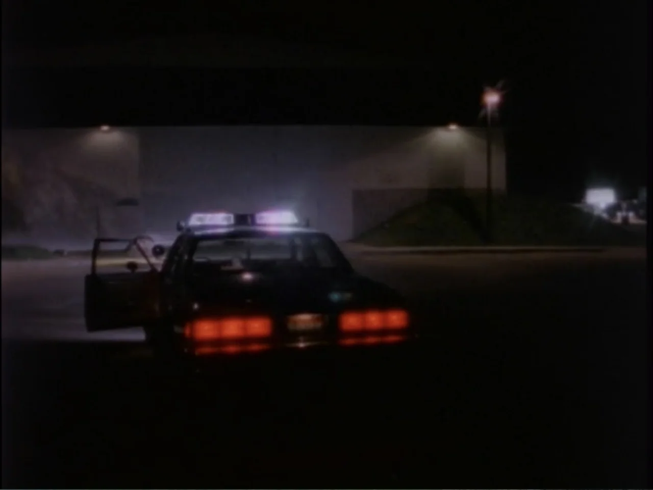 5 American Handguns - 5 American Kids (1995) movie still: wide — A dark, paved area at night with a large building structure in the distance. A…