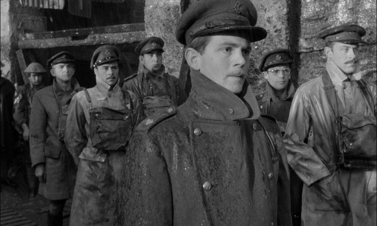 King & Country (1964) movie still: medium — A line of soldiers in military uniforms stands at an outdoor industrial docking area. It is raining and…