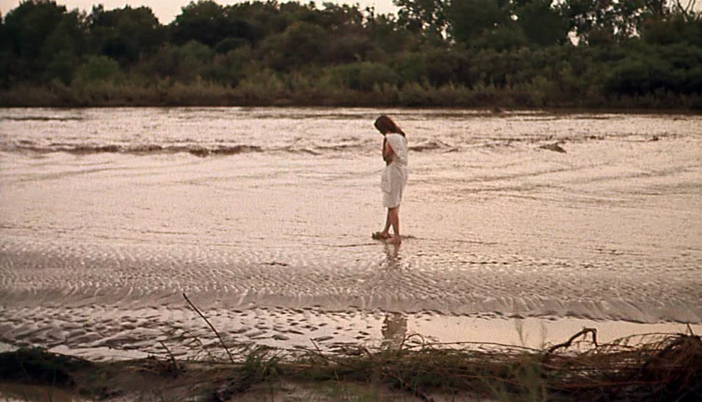 Badlands (1973) movie still: wide — A wide, shallow river flows through a rural landscape with dark treelines in background. A solitary woman in a…