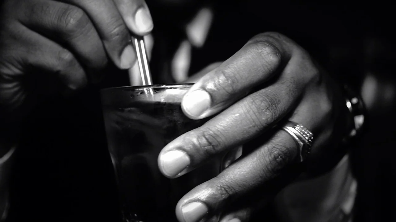 Kokomo City (2023) movie still: closeup — A close-up view shows a person's hand holding a glass and stirring it. A metal stirrer is being moved…