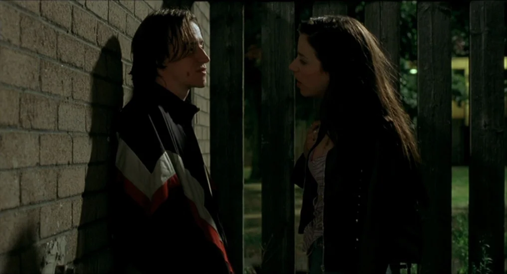 All or Nothing (2002) movie still: medium — A dark outdoor space flanked by a textured brick wall and wooden fence slats. A man and a woman stand…
