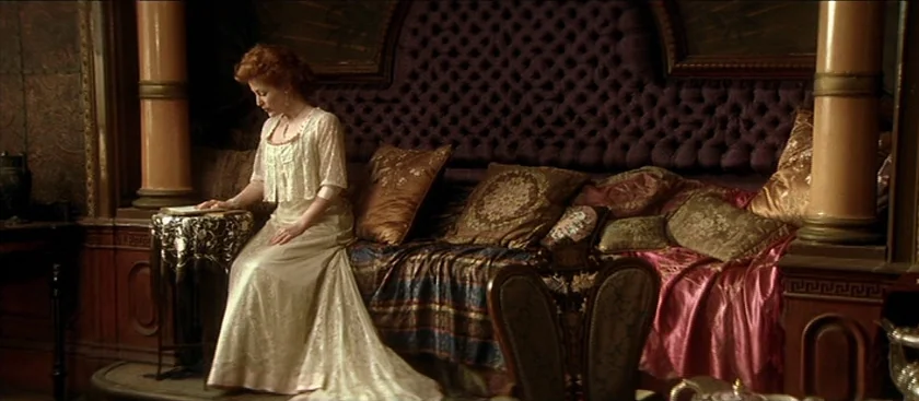 The House of Mirth (2000) movie still: fullbody — An opulent sitting room featuring a large tufted purple velvet daybed and antique columns. A woman…