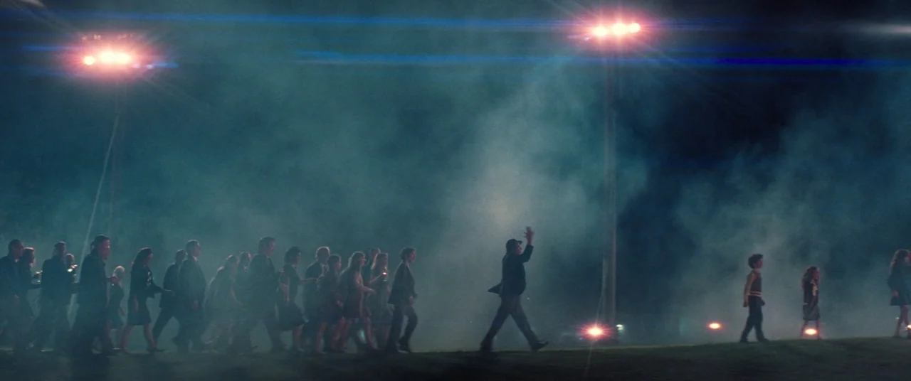 Licorice Pizza (2021) movie still: wide — A large crowd of people moves across a dark, fog-covered grassy field at night. Two tall stadium light…