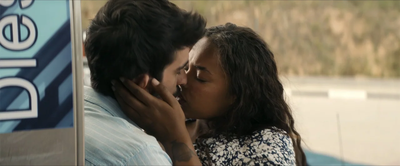 A Hipster in Rural Spain (2024) movie still: closeup — A pair of individuals kiss at a gas station near a blue metal pump. The man and woman embrace…