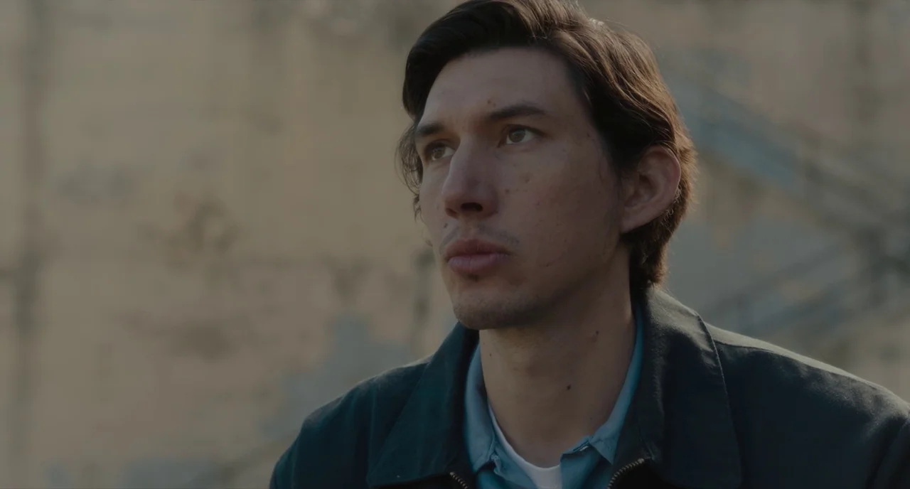Paterson (2016) movie still: closeup — A man stands outdoors against a weathered beige wall with visible concrete texture. He gazes forward while…