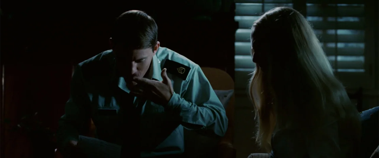 Dear John (2010) movie still: medium — A dark indoor space features a soldier reading a letter by window light. A blonde woman sits in the adjacent…