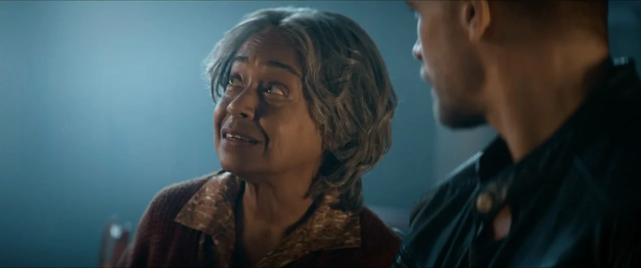 Man with No Past (2025) movie still: medium — A dimly lit indoor space with a cool blue color cast provides a quiet setting. An older woman in a…