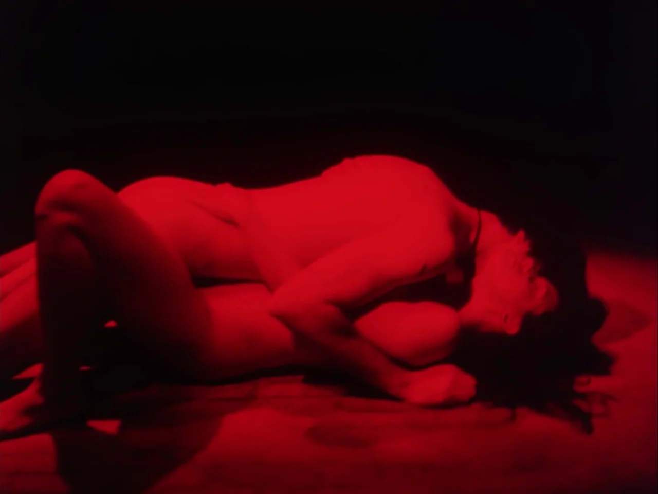 Subconscious Cruelty (2000) movie still: medium — Two individuals lie together on a dark floor in a dimly lit room. They are tightly embraced, their…