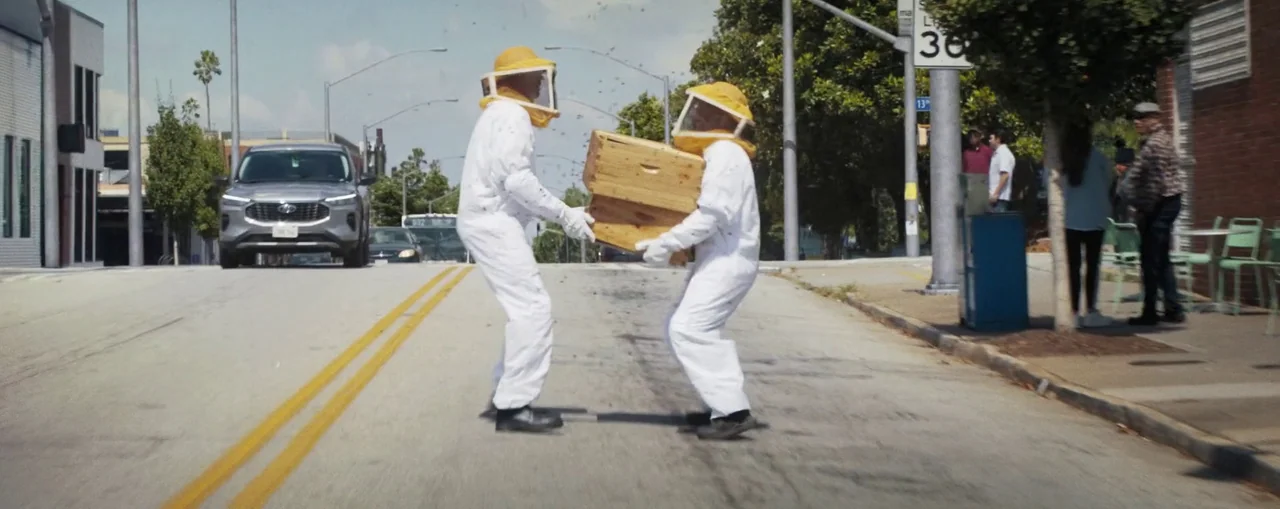 The Naked Gun (2025) movie still: fullbody — Two individuals wearing full protective white beekeeping suits walk across a paved city street. They…
