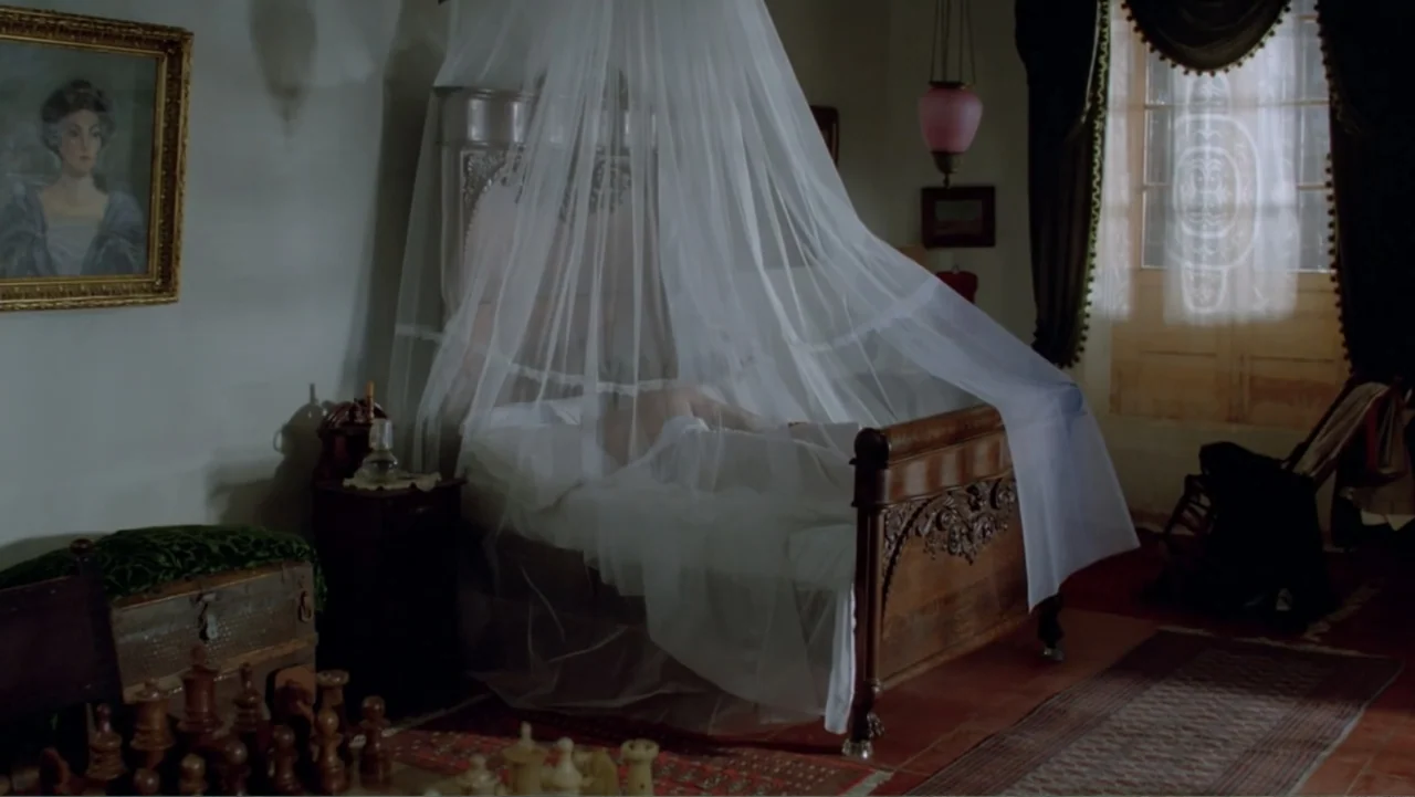 Toña Machetes (1985) movie still: wide — A 19th-century bedroom features a dark wooden bed covered by a translucent white canopy. A chess set sits…