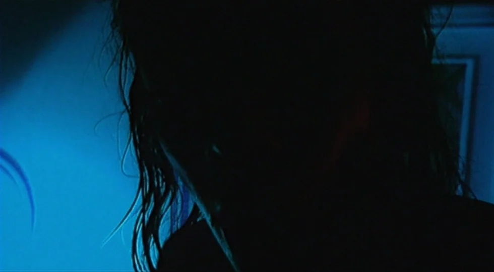 Scarlet Diva (2000) movie still: closeup — An interior space is cast in darkness with a vivid blue light source. A silhouette of a person appears in…