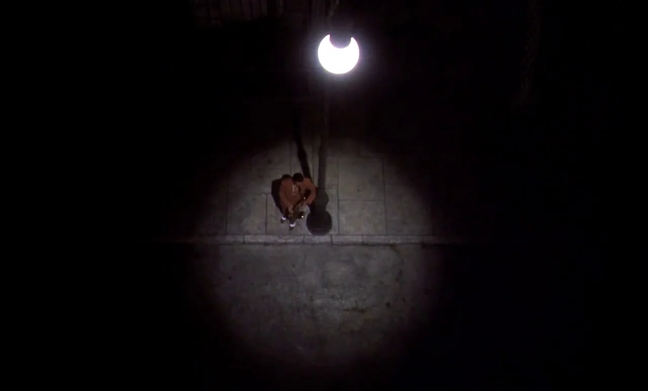 New York, New York (1977) movie still: wide — An overhead view looks down at a paved sidewalk at night. A lone man stands in a circle of light…