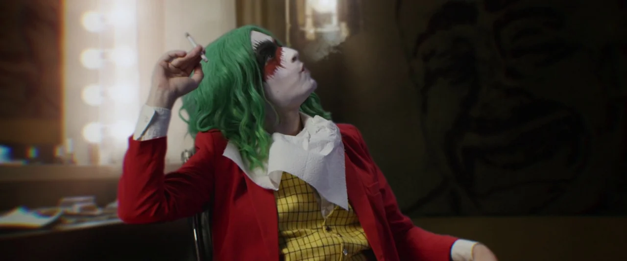 The People’s Joker (2022) movie still: medium — A dimly lit makeup room features vanity lights and a dark wall with a drawing. A person dressed as a…