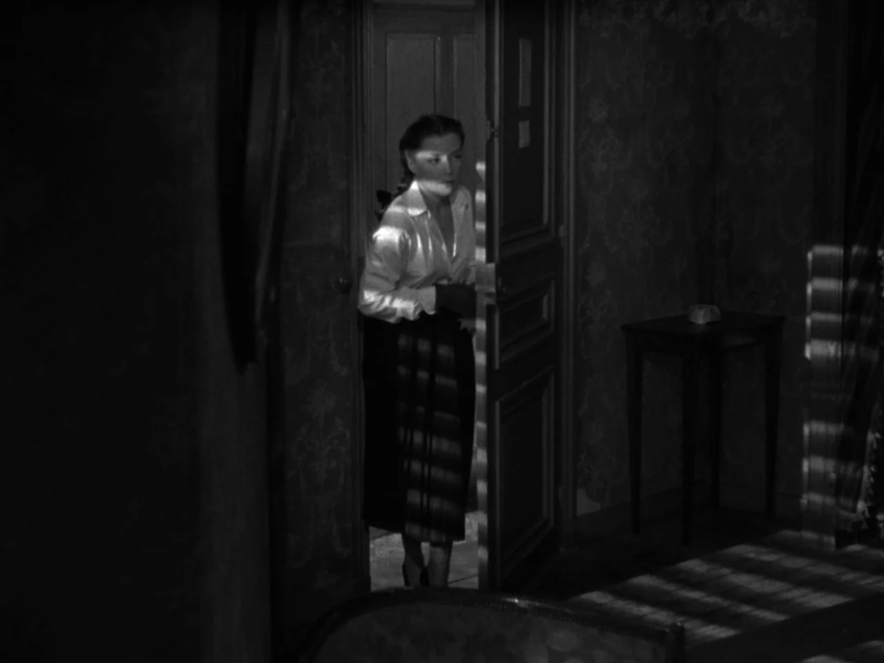 Diabolique (1955) movie still: fullbody — A dark space features a woman standing in a narrow, partially opened wooden doorway. She stands in the…