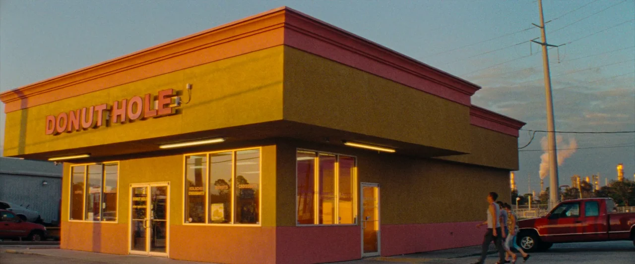 Red Rocket (2021) movie still: wide — A stucco building with pink and mustard yellow walls stands at a paved corner. Two people walk past the front…