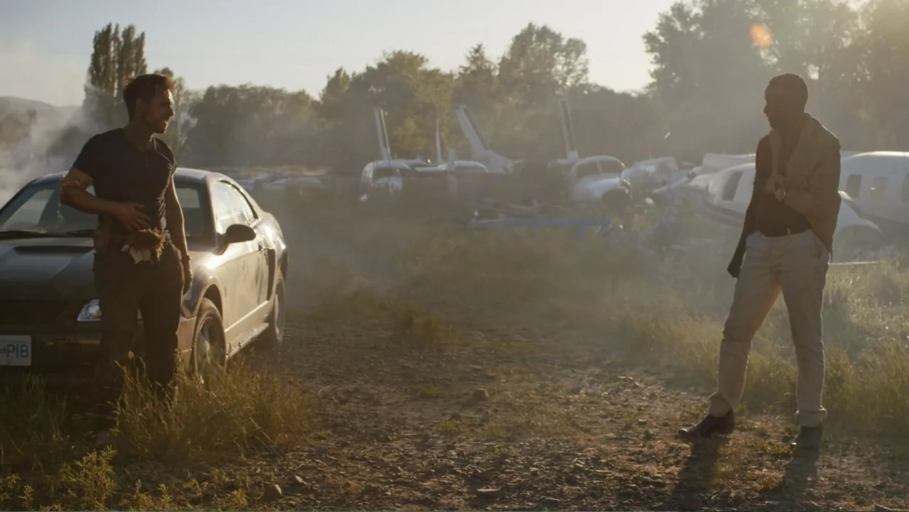 Bullet Proof (2022) movie still: fullbody — A dusty outdoor clearing filled with discarded aircraft wreckage and overgrown grass at sunset. Two men…