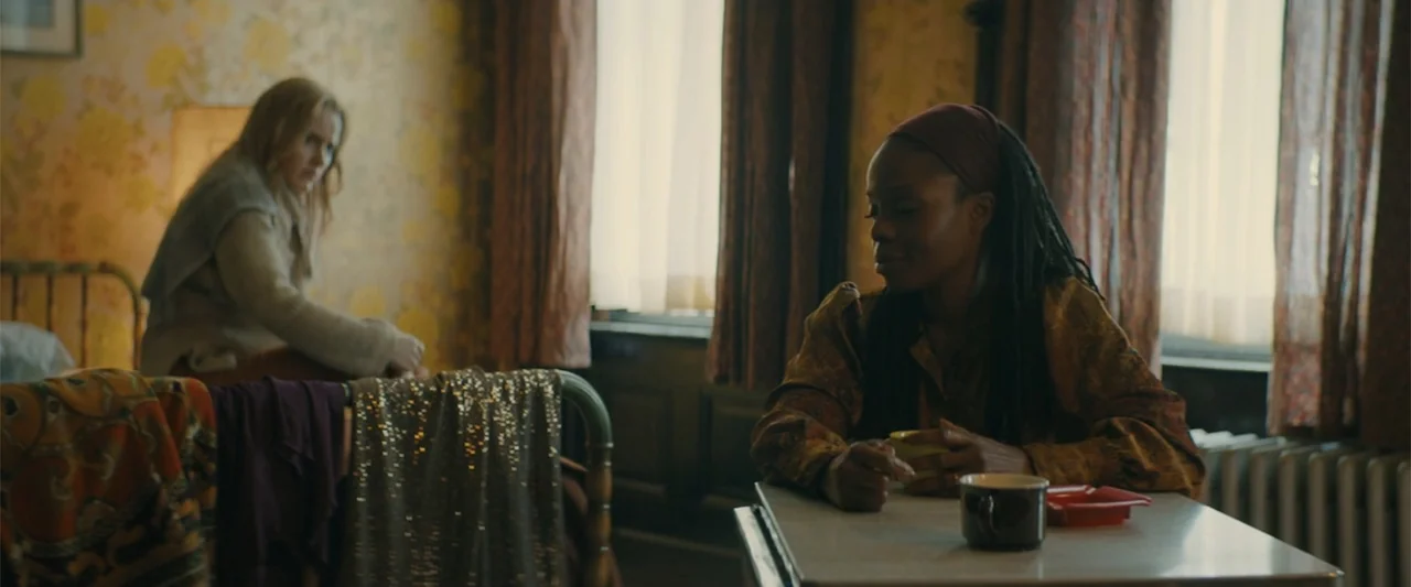 I'm Your Woman (2020) movie still: medium — A bedroom with yellow floral wallpaper features a metal-framed bed and a small table. One woman sits at…