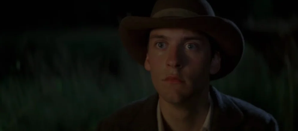 Ride With The Devil movie still: closeup — A man stands outdoors in a dark field at night with foliage visible nearby. He is illuminated by dim,…