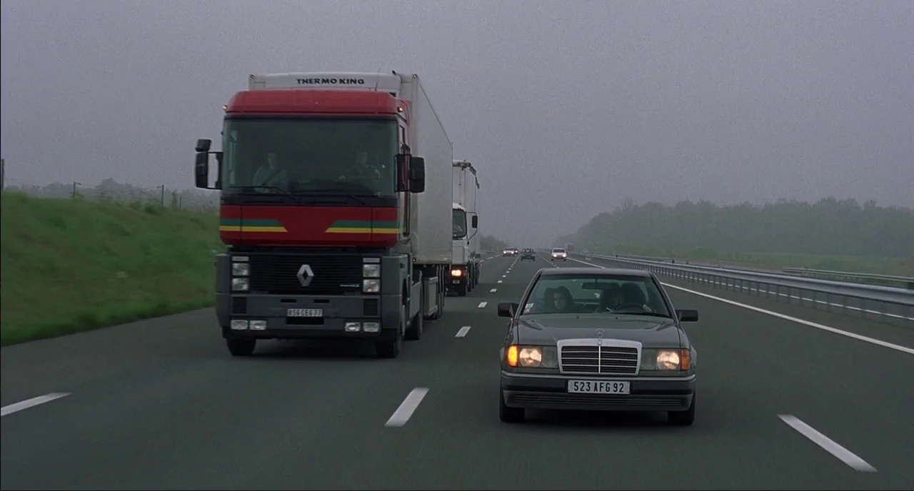Fat Girl (2001) movie still: wide — A grey Mercedes sedan travels along a multi-lane highway near a large freight truck. The road surface is grey…