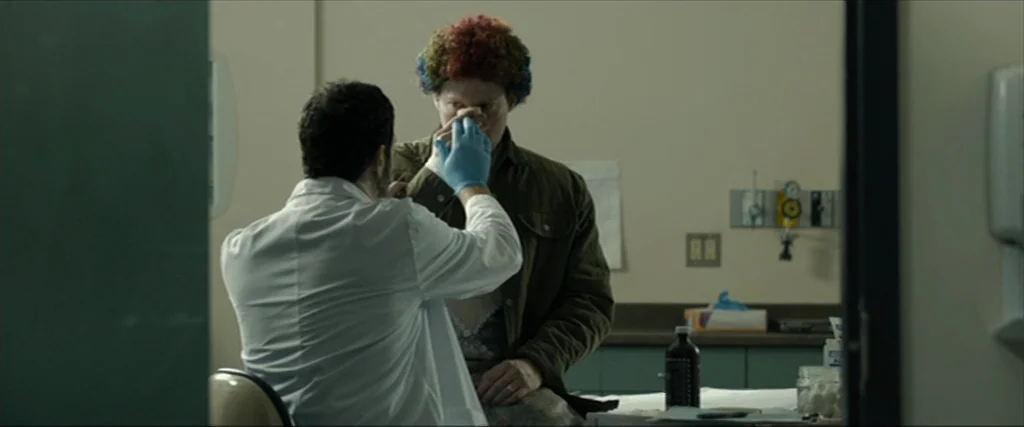 Clown (2014) movie still: medium — A clinical examination room with beige walls and medical equipment mounted on the wall. A doctor in a lab coat…