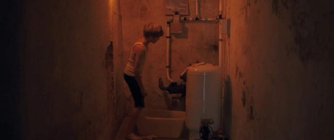 Goodnight Mommy (2014) movie still: medium — A basement room features textured, peeling walls and exposed plumbing pipes. A young boy stands by a…