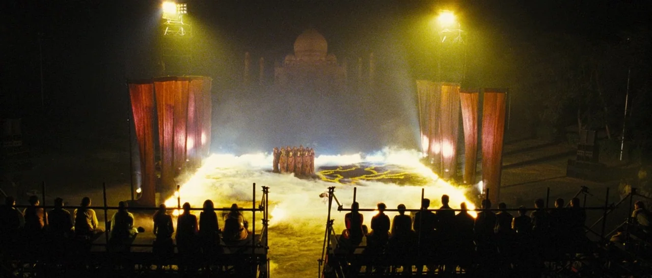 Slumdog Millionaire (2008) movie still: wide — An outdoor night performance space features a raised platform covered in thick white fog. A group of…