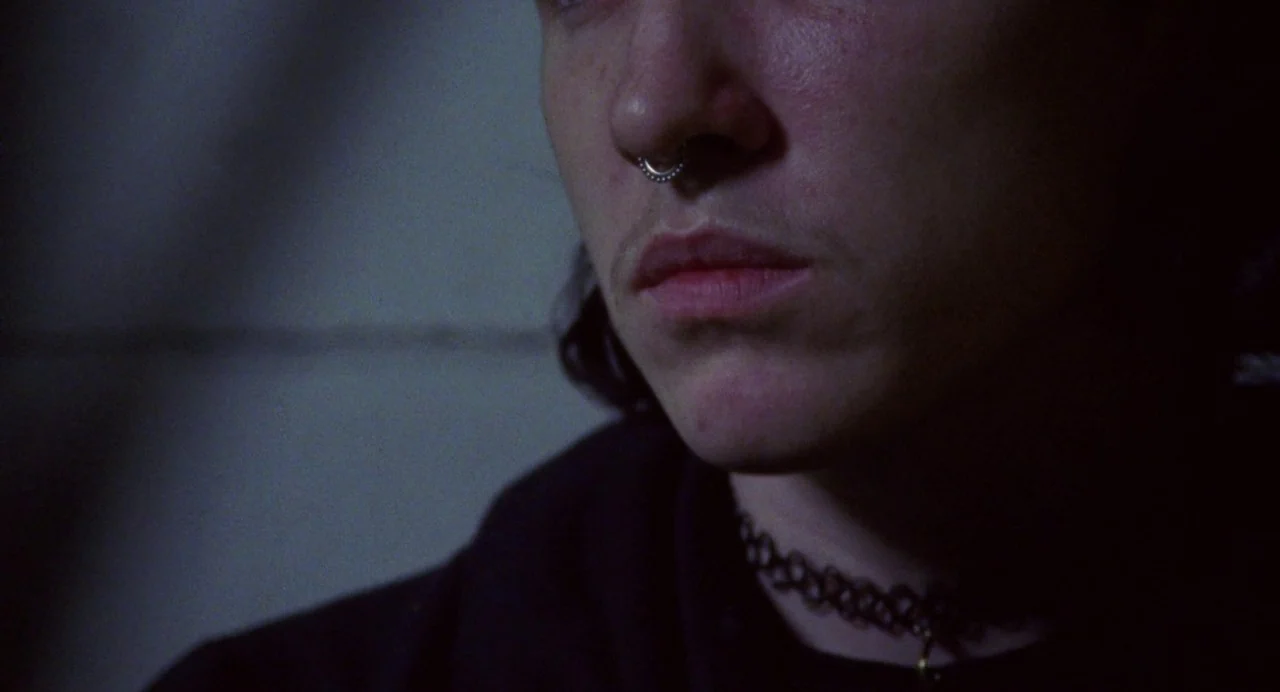 So Pretty (2019) movie still: closeup — A dimly lit indoor space with a visible seam on a textured wall surface. A person with dark hair and a…