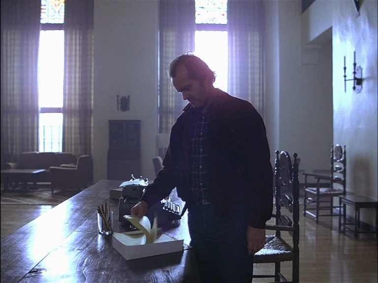 The Shining (1980) movie still: medium — A grand, sparsely furnished hall features large windows and high ceilings. A man stands at a long table,…