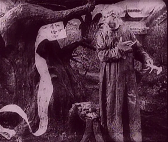 Destiny (1921) movie still: fullbody — A dense grove features a large, twisted tree with a paper scroll draped over it. An elderly, bearded man in a…