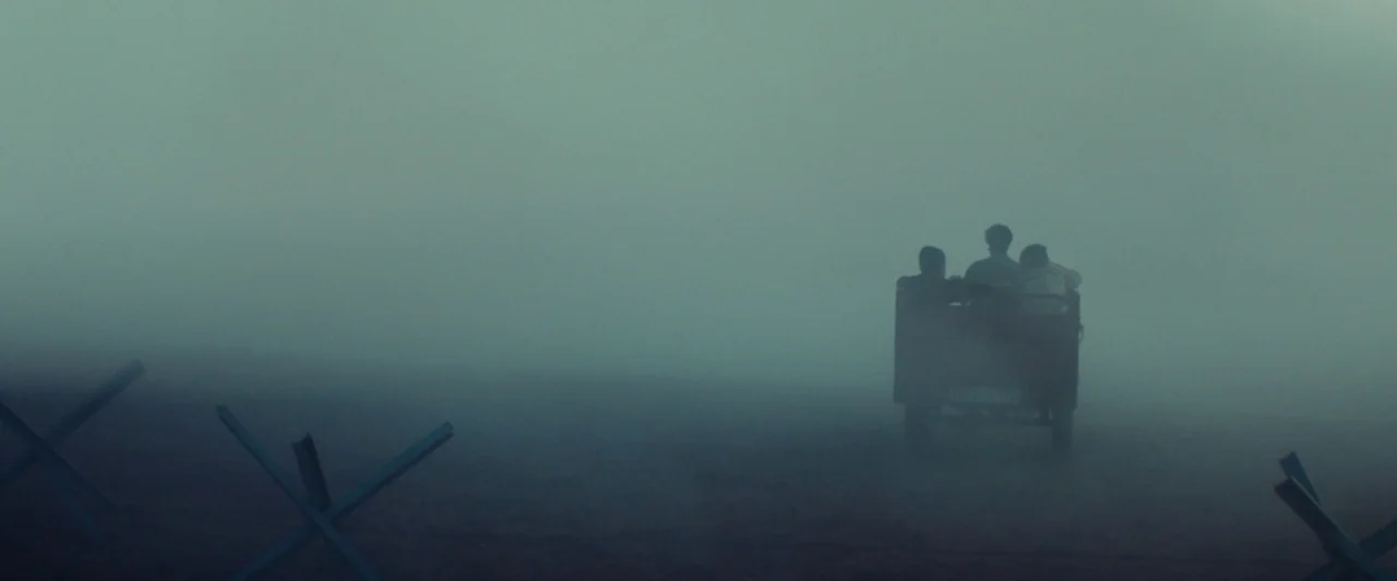 Animalia (2023) movie still: wide — A desolate, fog-covered landscape scattered with steel anti-tank obstacles. Three individuals in a small vehicle…