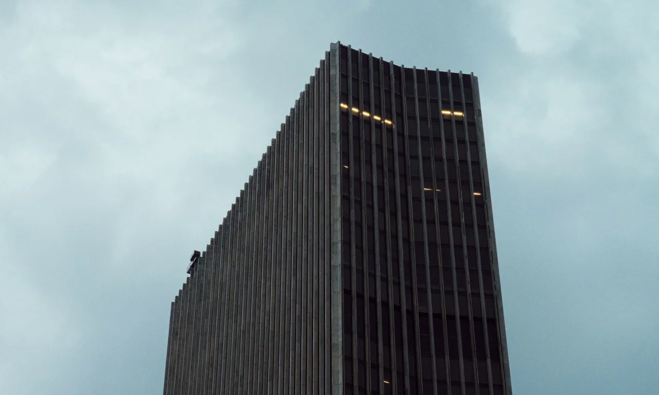 Resurrection (2022) movie still: establishing — A tall, monolithic skyscraper with vertical concrete ribs rises against a clouded grey sky. Several…