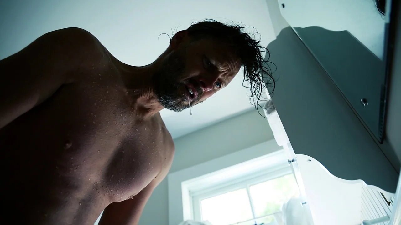 The Deeper You Dig (2019) movie still: medium — A sterile bathroom features white surfaces and a window partially obscured by cabinets. A shirtless…