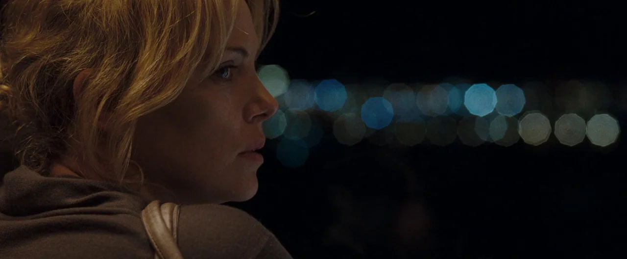 The Burning Plain (2008) movie still: closeup — A woman stands in the darkness at night overlooking a distant city. Blurred points of light from an…