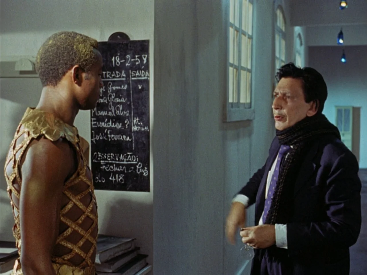 Black Orpheus (1959) movie still: medium — A man in a golden costume stands before a chalkboard with handwritten text. An older man in a suit…