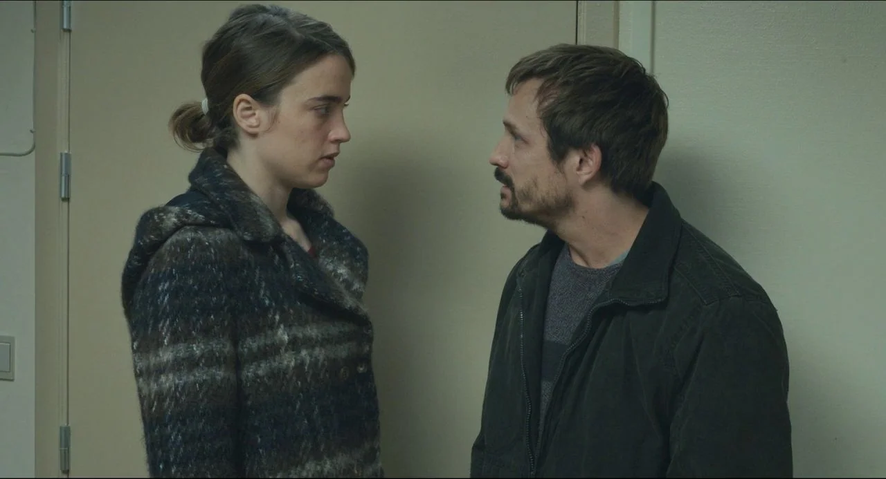 The Unknown Girl (2016) movie still: medium — A featureless hallway with beige walls and a single door frame behind them. A woman and a man stand…
