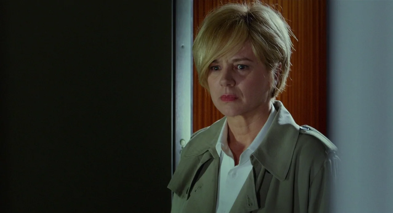 Julieta (2016) movie still: medium — A dimly lit indoor corridor with plain walls and a dark vertical frame. A blonde woman in a tan trench coat…