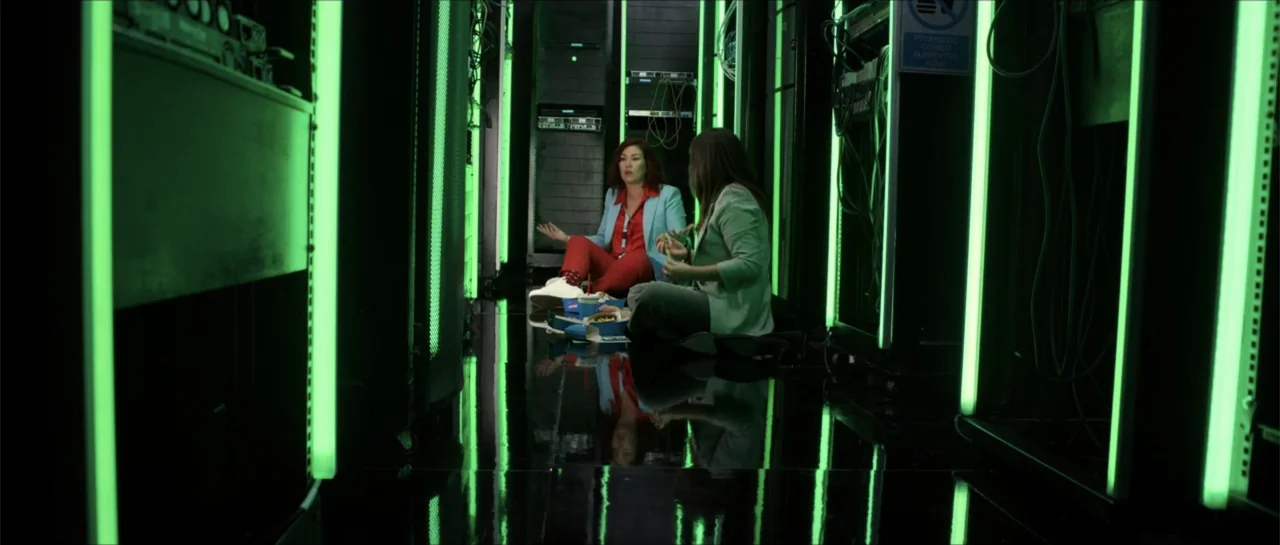 Disconnected 2 (2025) movie still: fullbody — Two women sit on the floor between rows of glowing green computer server racks. They are eating fast…